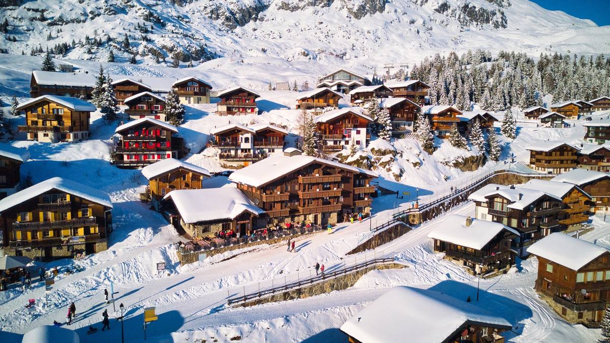 Aerial view of Bettmeralp's village center under fresh snow, timber chalets and a restaurant terrace lining a car-free snowy path with the ski slope and mountains behind