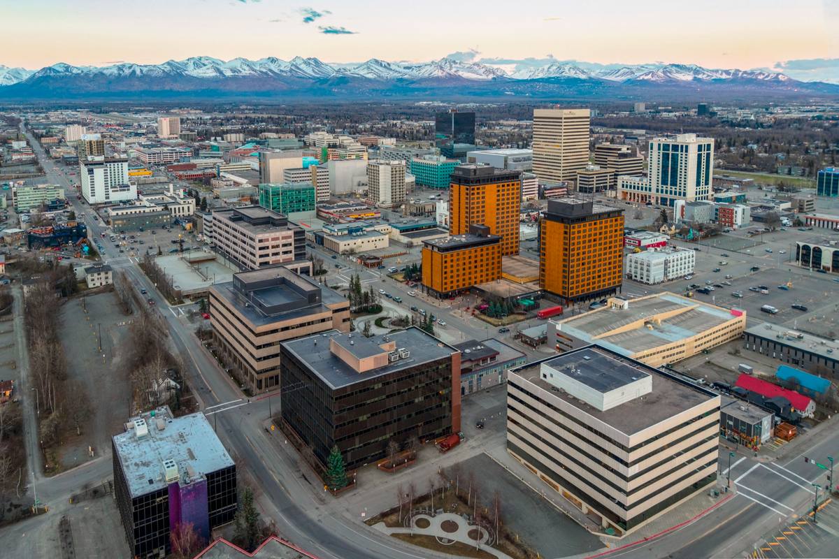An aerial view of downtown Anchorage's high-rise skyline in evening light, with the snow-capped Chugach Mountains stretching across the horizon behind the city