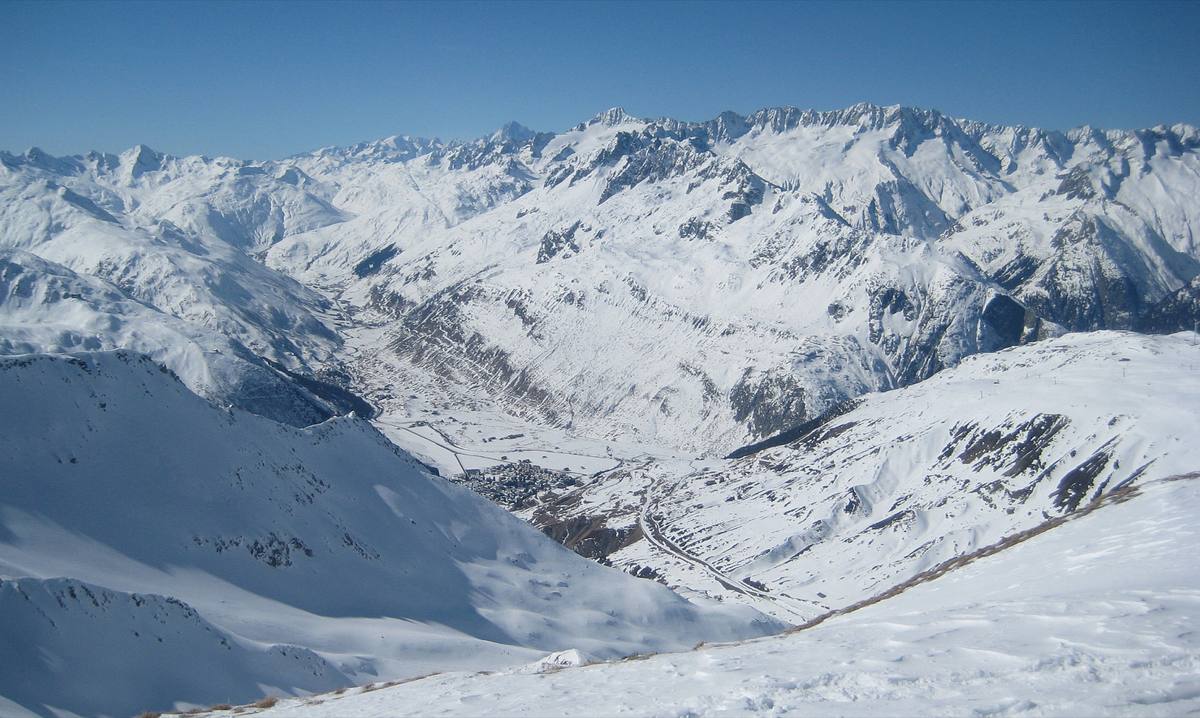 Winter panorama from the Pazolastock above the Oberalp Pass, looking down on Andermatt village on the snow-covered floor of the Ursern Valley, ringed by high Alpine peaks under a blue sky