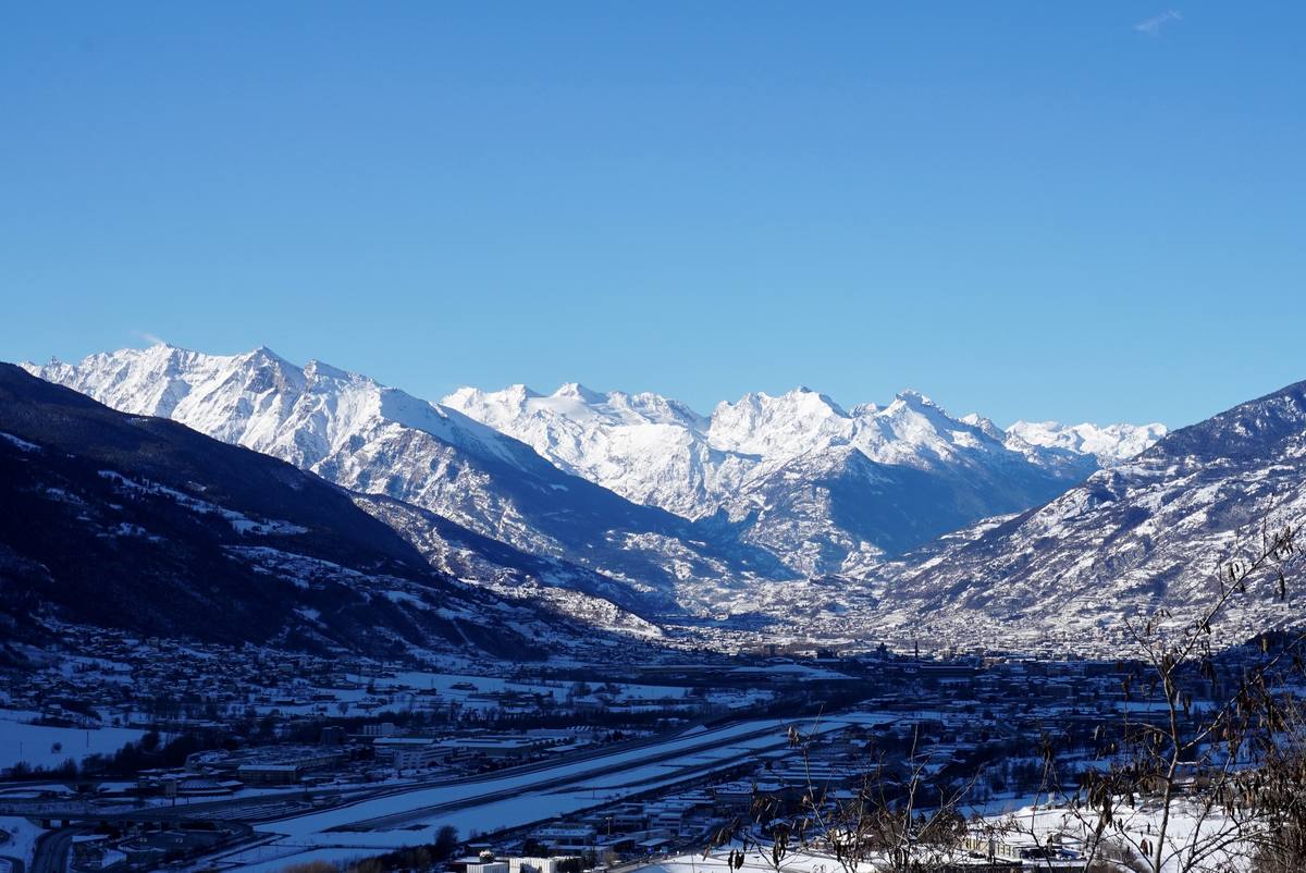 Winter view down the snow-covered Aosta valley, the city of Aosta spread across the flat valley floor beneath ranks of snow-capped peaks, with the airport runway strip in the foreground