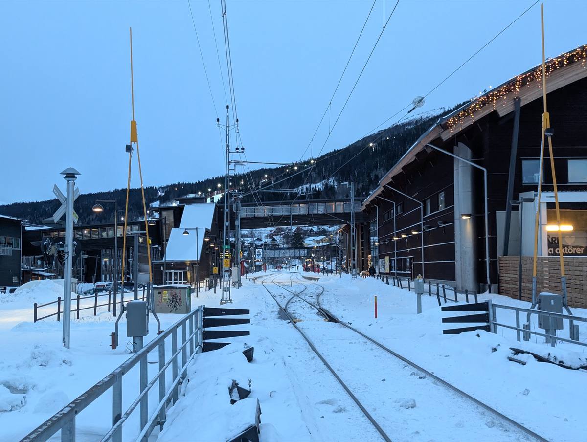 Snow-covered railway tracks curve past the Åre station buildings at dusk, string lights along the eaves and a floodlit ski slope on the mountainside behind