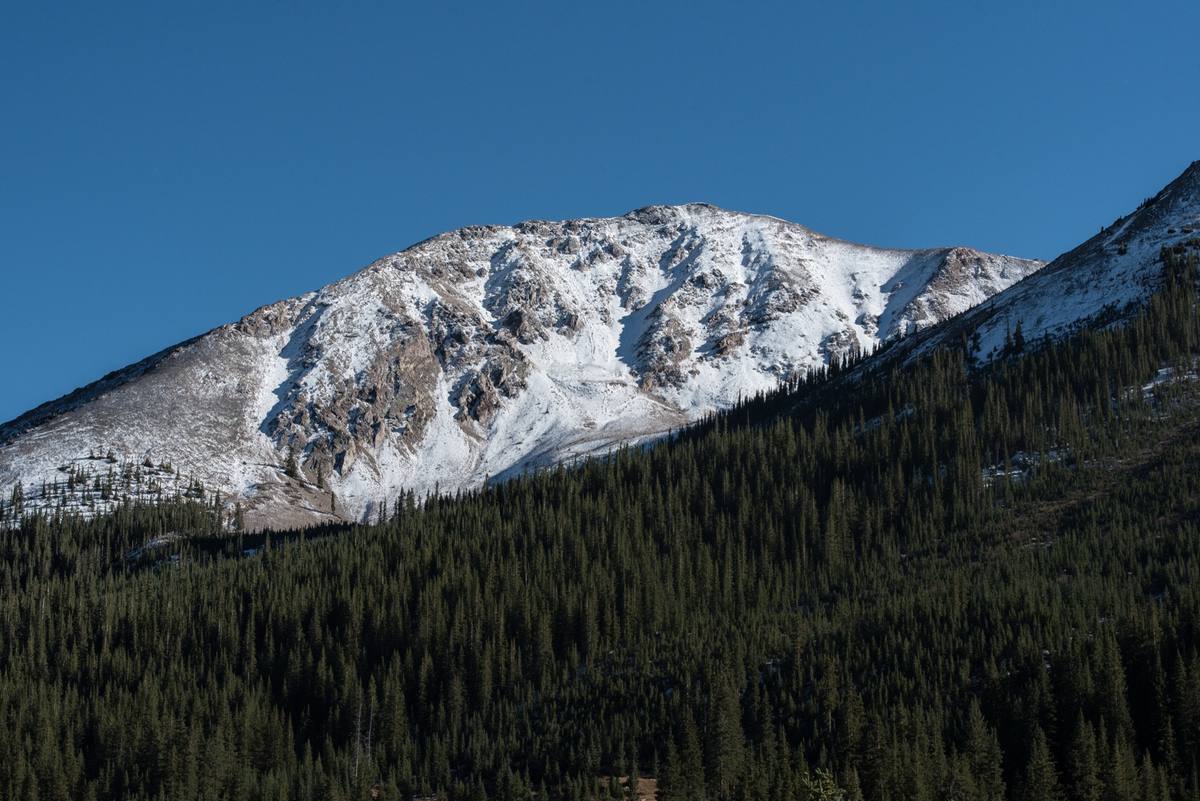 A snow-covered rocky summit above a dense dark conifer forest under a deep blue winter sky near Aspen, Colorado