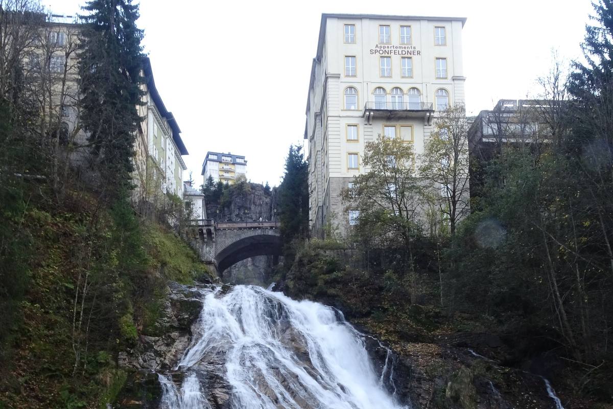 The Appartements Sponfeldner building rises on a cliff above a stone bridge, with the Gasteiner Ache waterfall cascading through the gorge below in central Bad Gastein