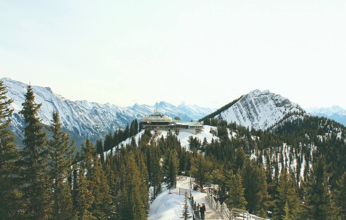 A mountaintop summit station with an observation deck and a Canadian flag on a snow-covered ridge, a boardwalk and snowy conifers in the foreground and range after range of snow-covered Rockies behind