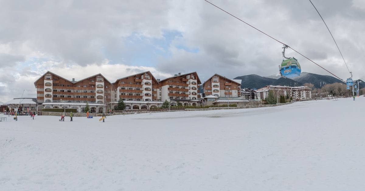 A Bansko-branded gondola cabin rides its cable over a snow-covered slope at the edge of town, where children sled below the timber-balconied hotels of the gondola quarter and forested Pirin foothills rise into cloud behind