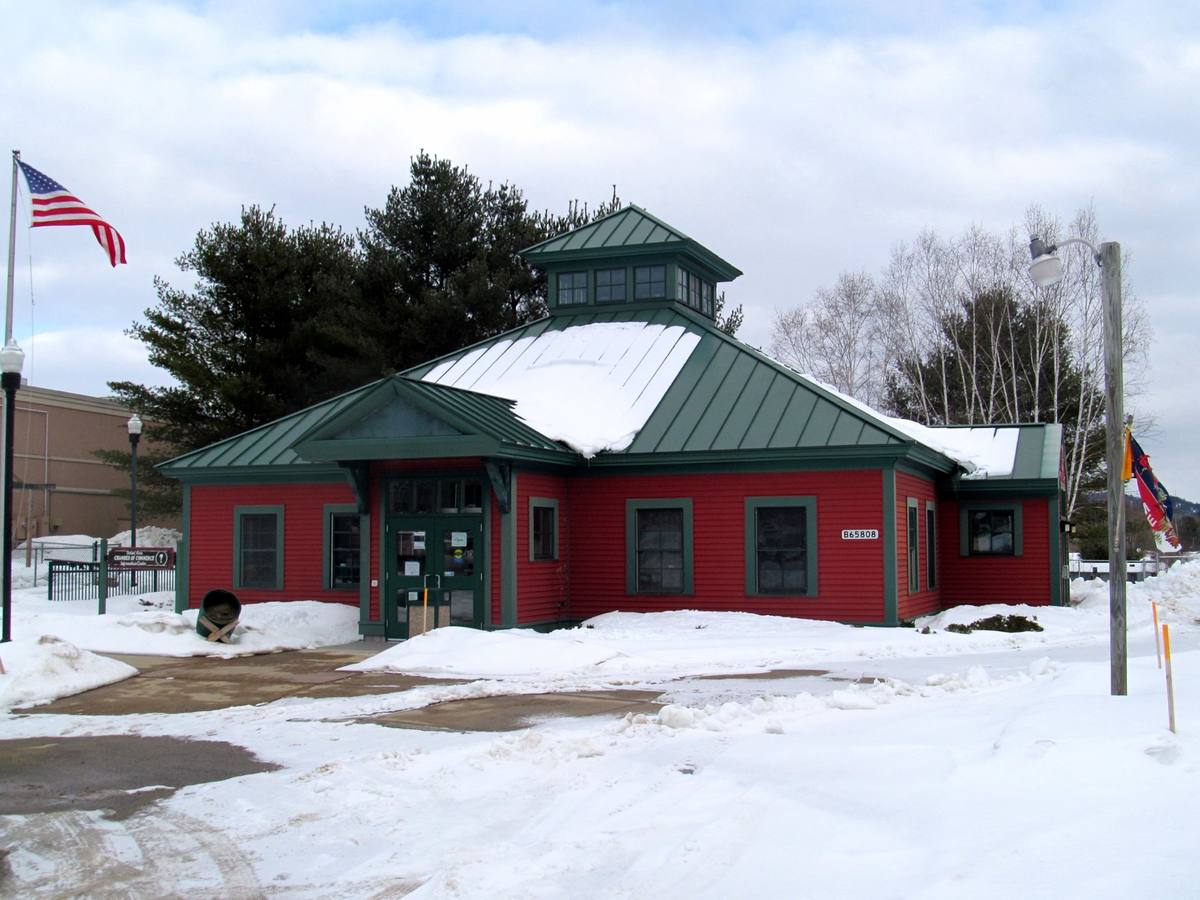 The red clapboard former Bethel station, now the chamber of commerce, under deep midwinter snow with a US flag flying and plowed snowbanks across the lot
