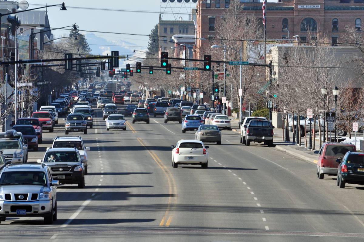 Traffic on Bozeman's Main Street on a winter afternoon, brick storefronts and "The Bozeman" building lining the block with snow-dusted rooflines and hazy mountains beyond