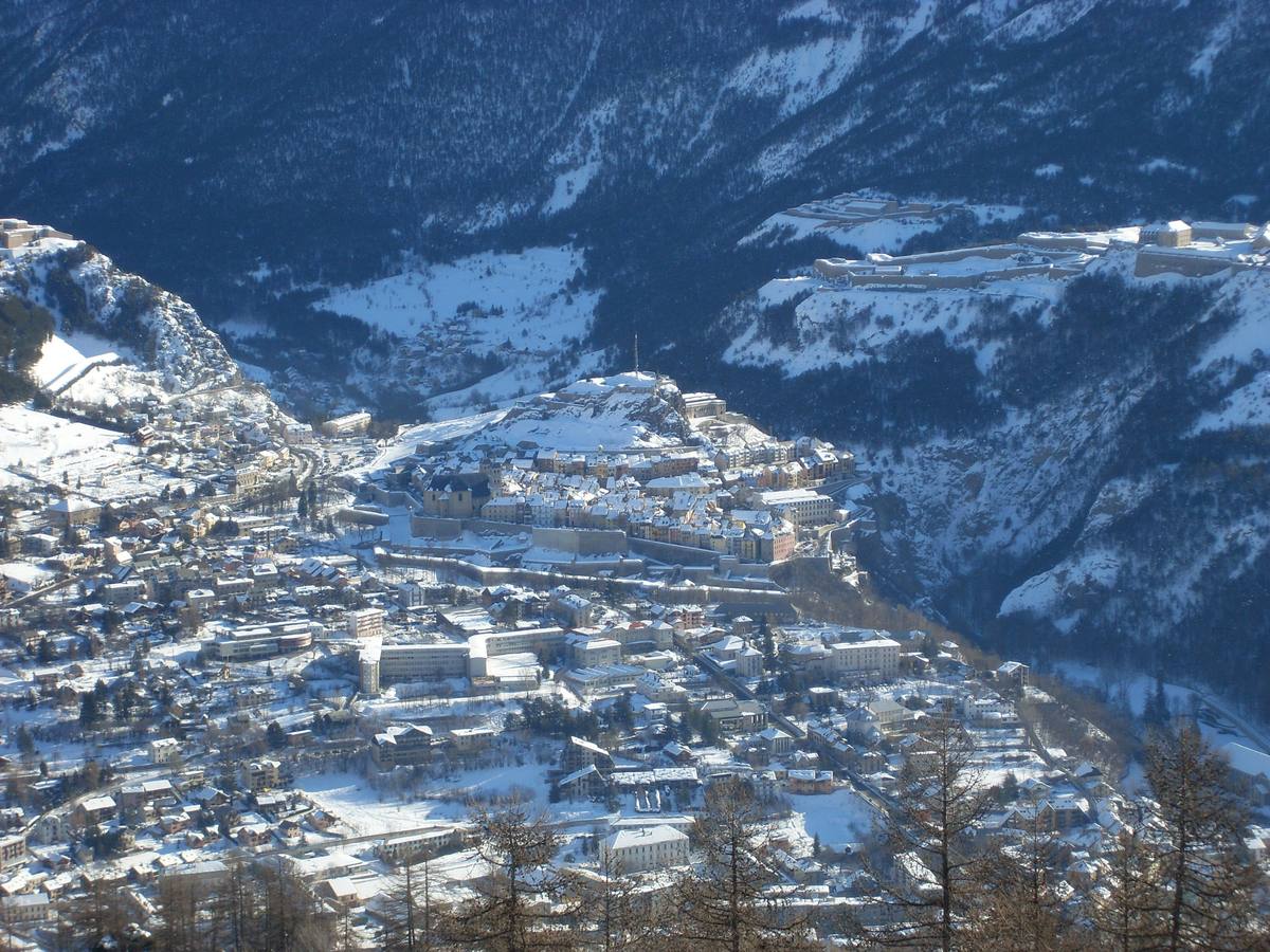 Briançon under deep snow seen from the slope above, the walled Cité Vauban on its rocky spur at centre with fort terraces on the plateau behind, and the lower town spreading across the white valley floor