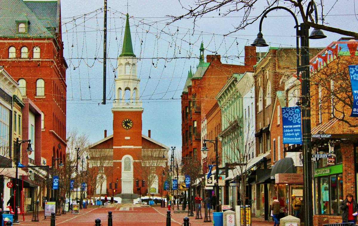The brick pedestrian Church Street Marketplace in winter, strings of lights above the street and snowbanks below the white steeple of the First Unitarian church