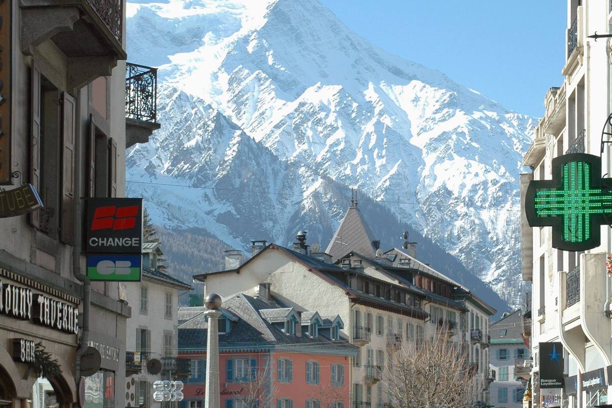 Chamonix's shopping street — taverne signs, a glowing pharmacy cross and pastel facades — beneath the snow-plastered face of the Aiguille du Goûter