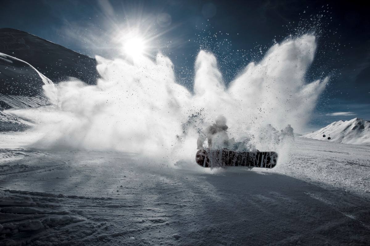A snowboarder carving hard on a Davos piste throws up a wall of powder against a deep blue sky, with gondola cabins and snow-covered peaks beyond