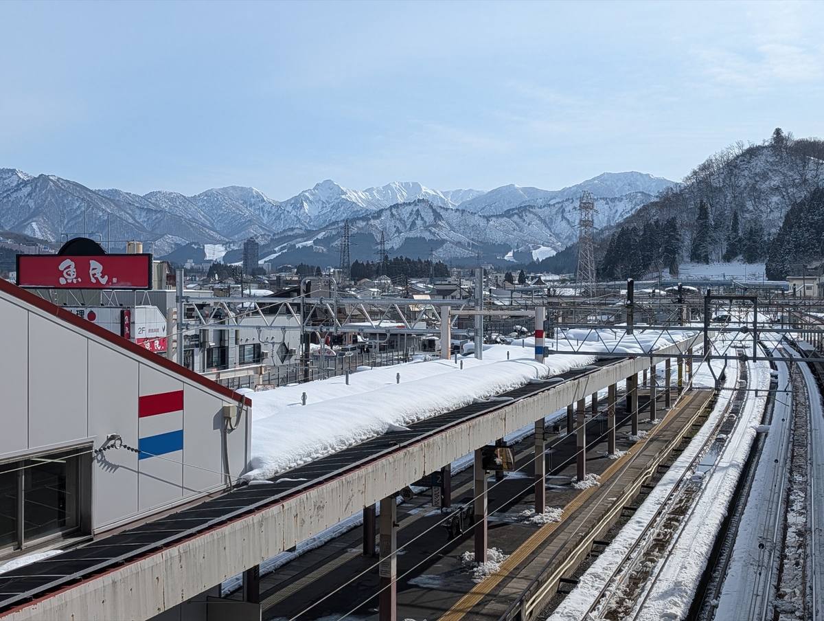 Deep snow banked on the platform canopies and tracks at Echigo-Yuzawa station, with the town's rooftops beyond and a wall of snow-covered mountains carrying pale ski runs behind