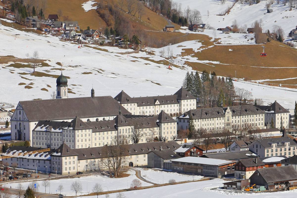 Kloster Engelberg's baroque monastery complex, its onion-domed clock tower rising above the roofline, sits under snow at the edge of town with a ski slope and cable car climbing the valley wall behind