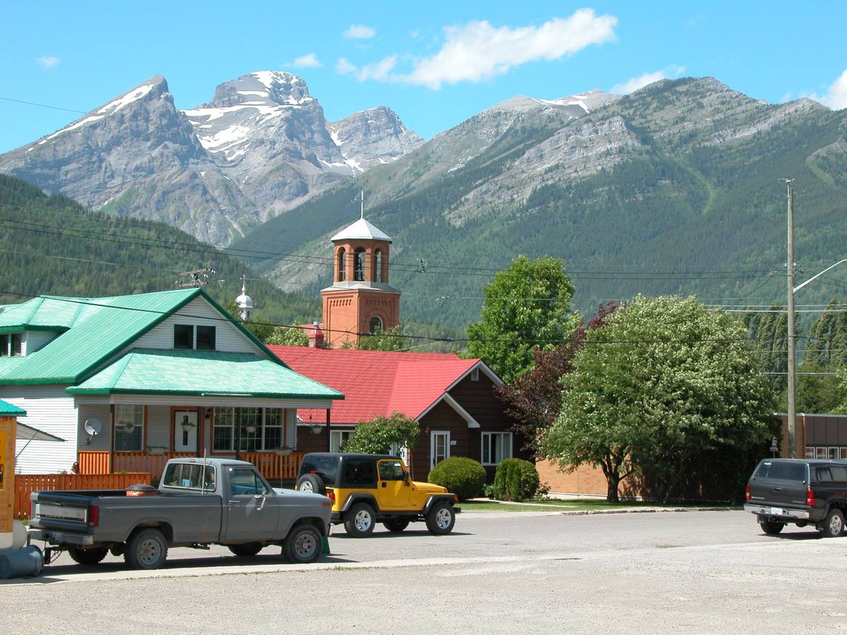 Houses with green and red metal roofs and parked pickup trucks in the foreground, a brick church bell tower behind them, and the snow-streaked peaks of the Three Sisters and Mount Proctor rising over downtown Fernie, British Columbia
