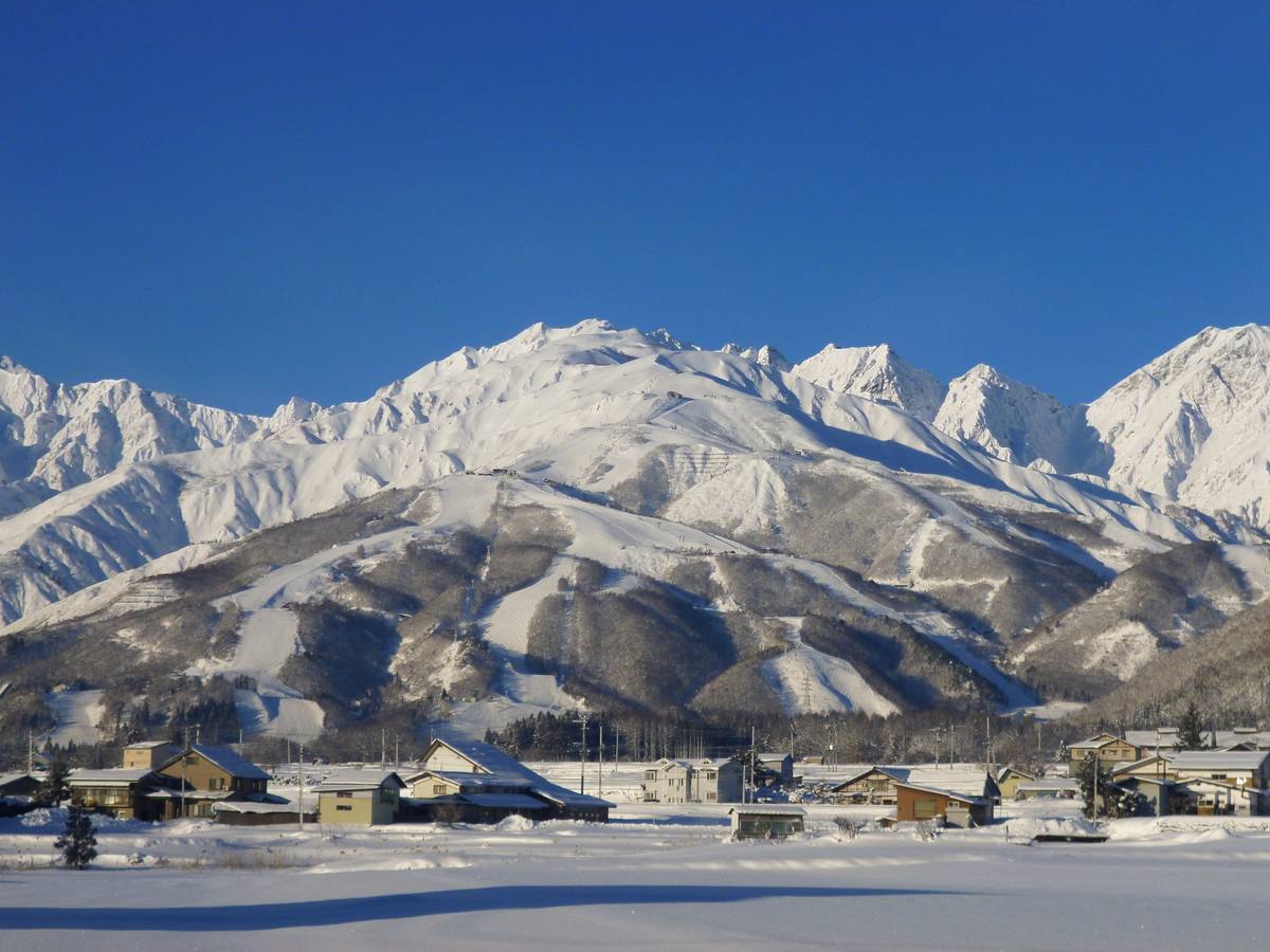 Snow-covered farmhouses and barns on the valley floor below the pisted ski runs of Happo-one, with the jagged Hakuba peaks rising white against a deep blue winter sky