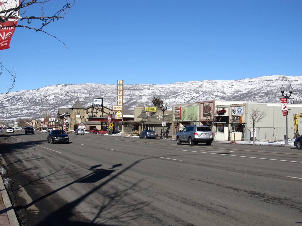 Heber City's wide Main Street in winter sun, with the Alpine Lodge sign, a Soldier Hollow tubing banner on a lamppost and the snow-covered Wasatch ridge rising behind the strip