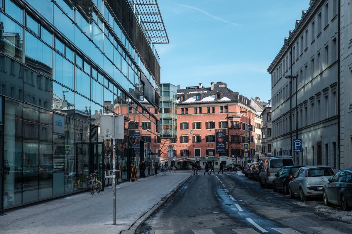 A central Innsbruck street on a clear winter day, snow lying on the mansard roof of the pink Hotel Schwarzer Adler and banked along the curb, pedestrians crossing between a glass-fronted modern building and a line of parked cars