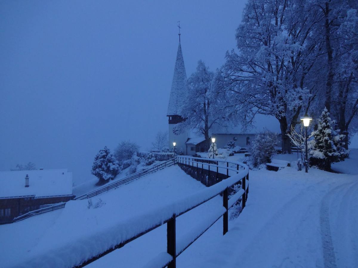 Wengen in heavy snowfall at dusk, the village church spire and snow-laden trees above a fenced path