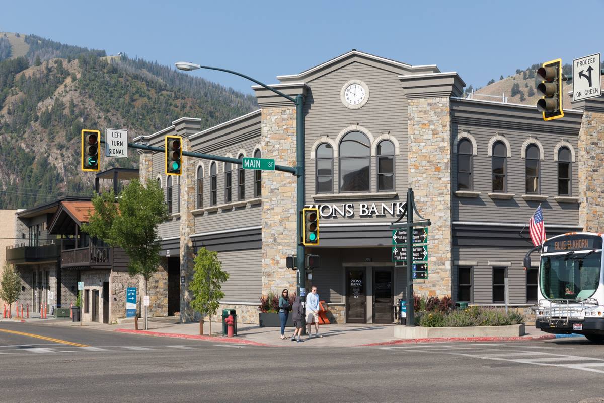 A Mountain Rides bus and pedestrians at Main Street and Sun Valley Road in downtown Ketchum, with signs to River Run, Warm Springs and Sun Valley Resort, and hills rising behind the corner bank building