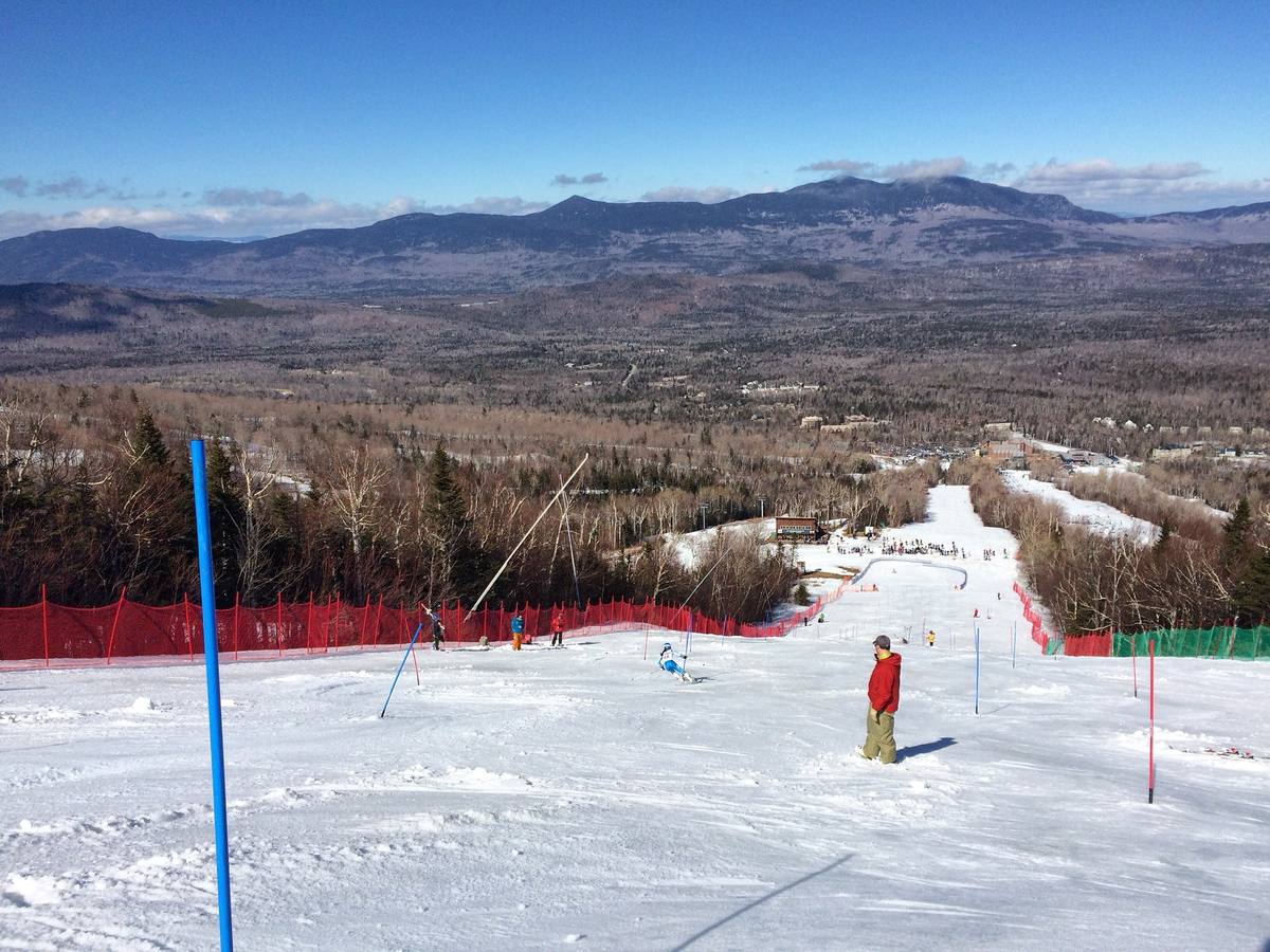 Skiers and race gates on a groomed snow trail high on Sugarloaf, looking down over the forested Carrabassett Valley to the base area and a long snow-streaked mountain range under a clear blue sky