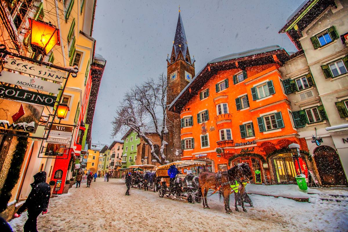Snow falls on Kitzbühel's pedestrianized Vorderstadt as a horse-drawn carriage waits before a row of painted gabled facades, with the Pfarrkirche St. Andreas clock tower rising behind