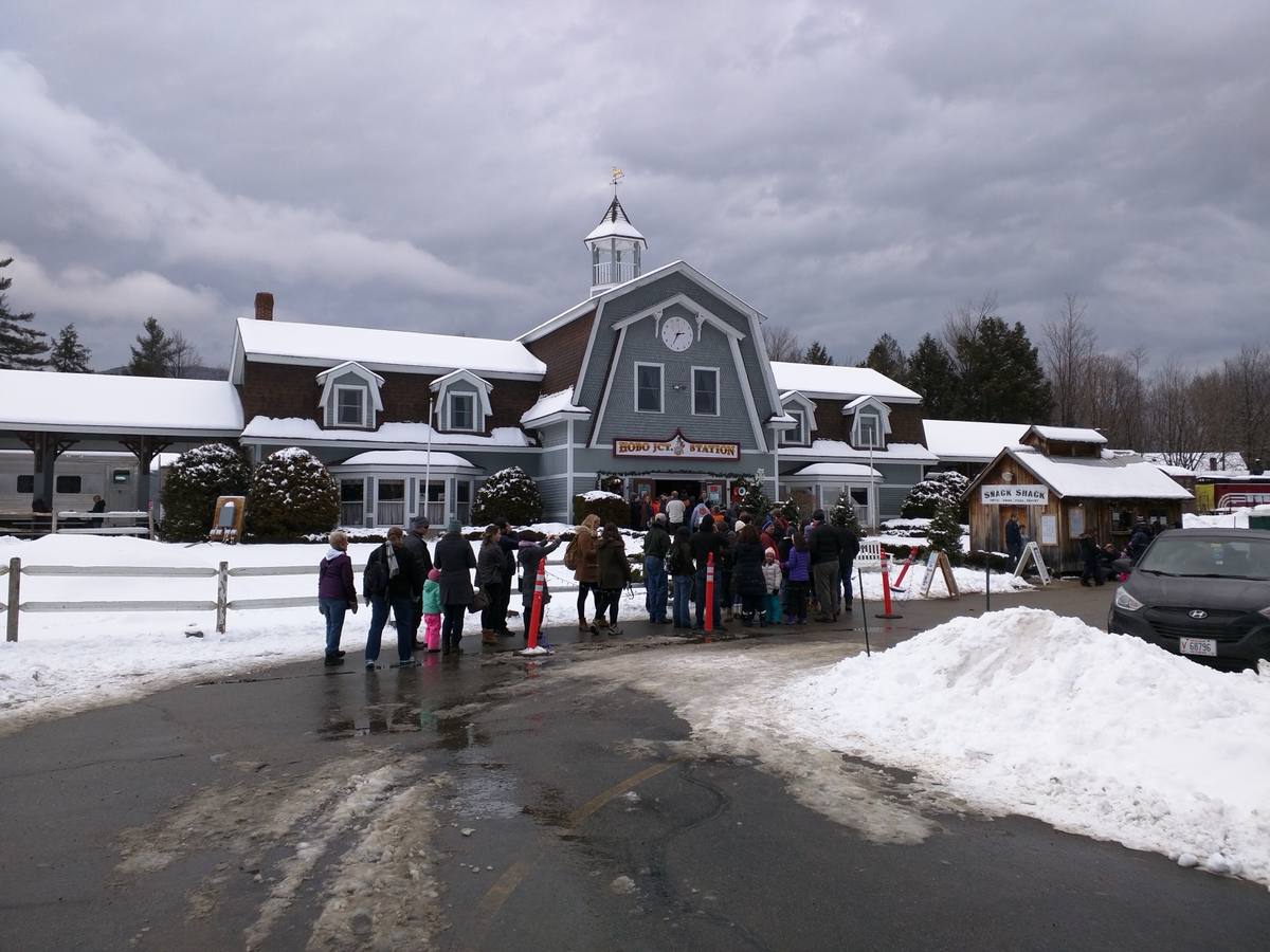 Snow-covered Hobo Junction station in Lincoln under heavy winter cloud, a line of people in parkas waiting at the door beside plowed snowbanks and a wet, salted road