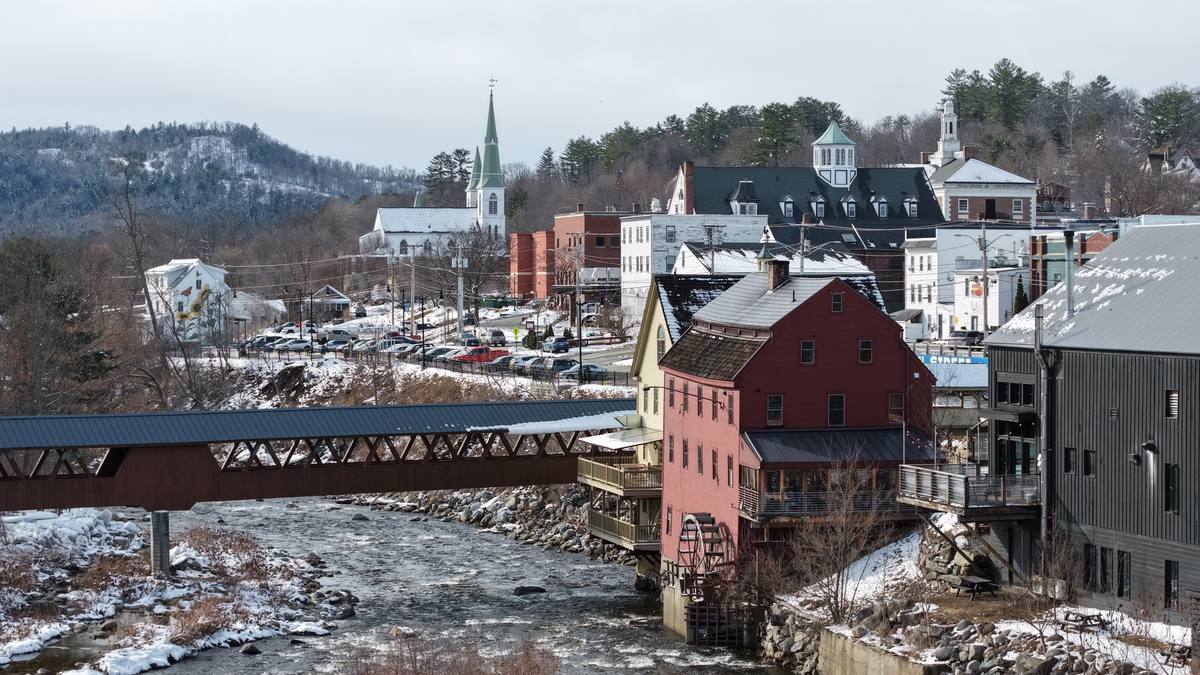 Snow-covered downtown Littleton above the Ammonoosuc River, with the red clapboard grist mill and its water wheel, a covered pedestrian bridge over the rapids, and a white church spire among Main Street rooftops