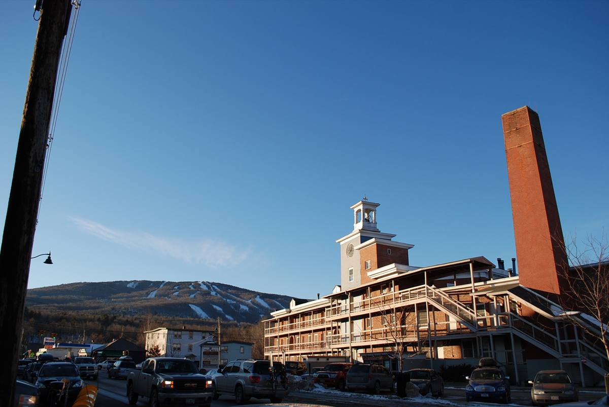 Snowy main street below the red-brick Mill clock tower and smokestack in Ludlow, with Okemo's white ski trails cutting through the wooded ridge behind under a clear winter sky