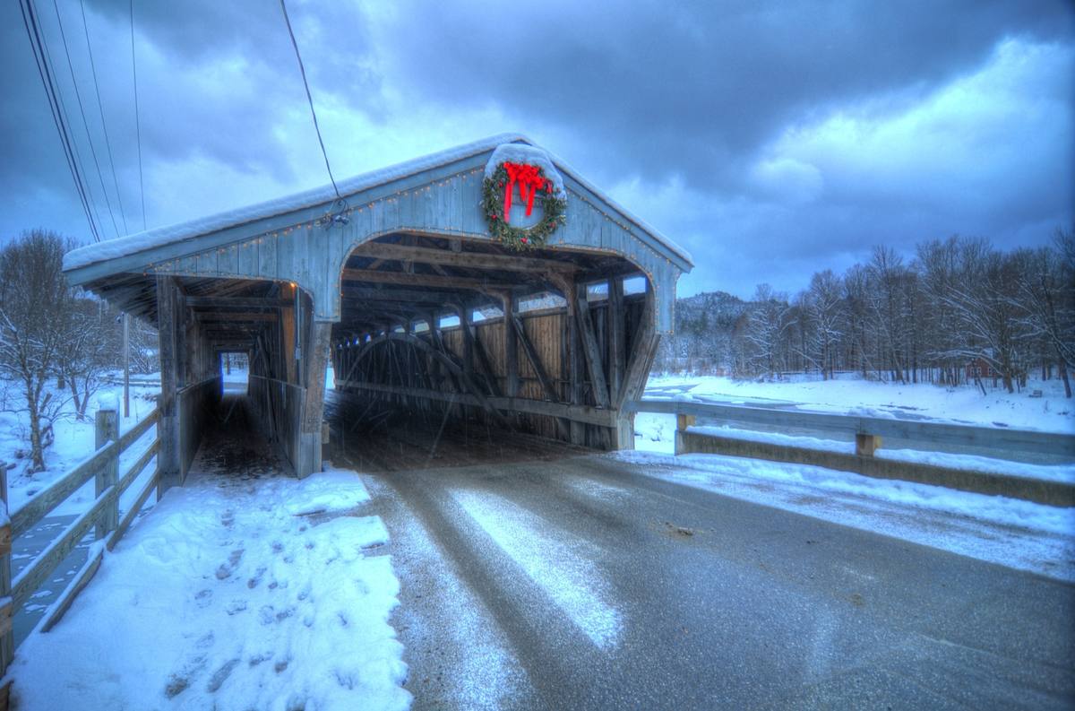 A snow-covered wooden covered bridge hung with a red-ribboned holiday wreath and string lights, its plowed road running through deep snowbanks beside a frozen river at dusk
