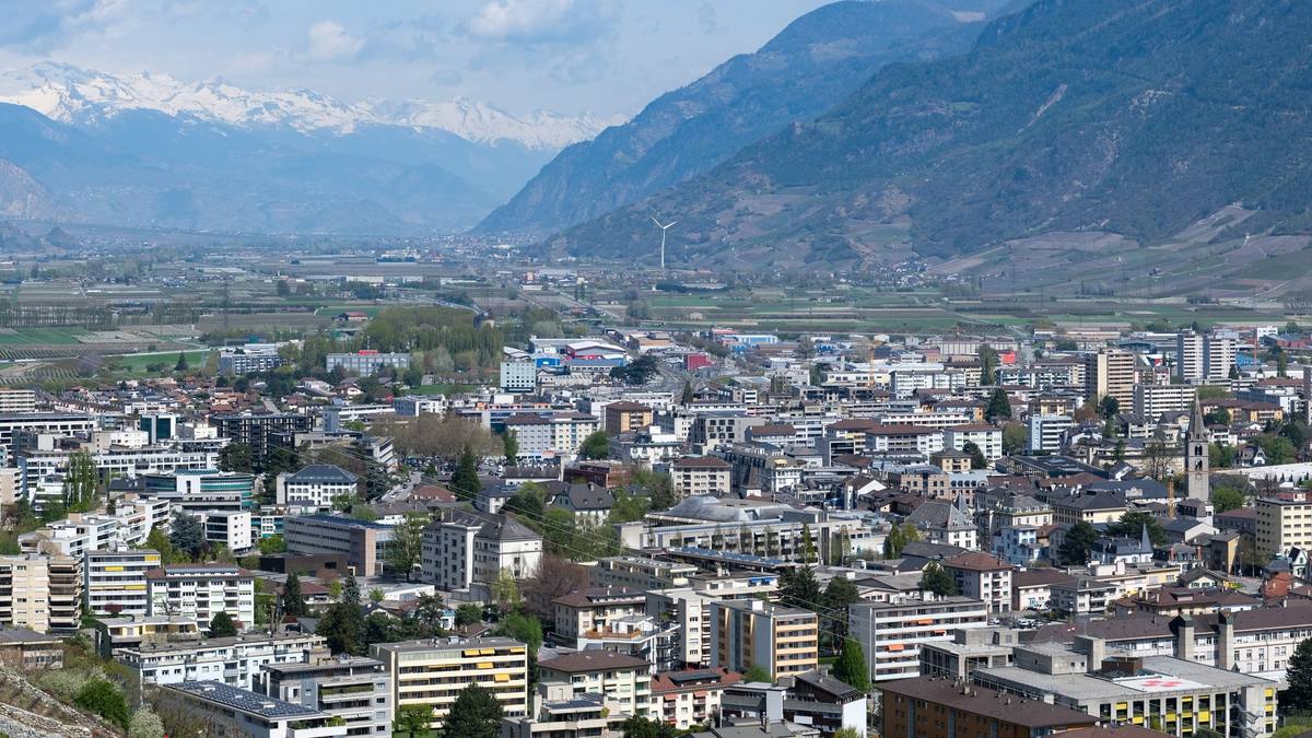 Martigny's town center seen from the hillside above it in April, apartment blocks and the church tower filling the flat Rhône valley floor, with snow-covered peaks closing the valley beyond