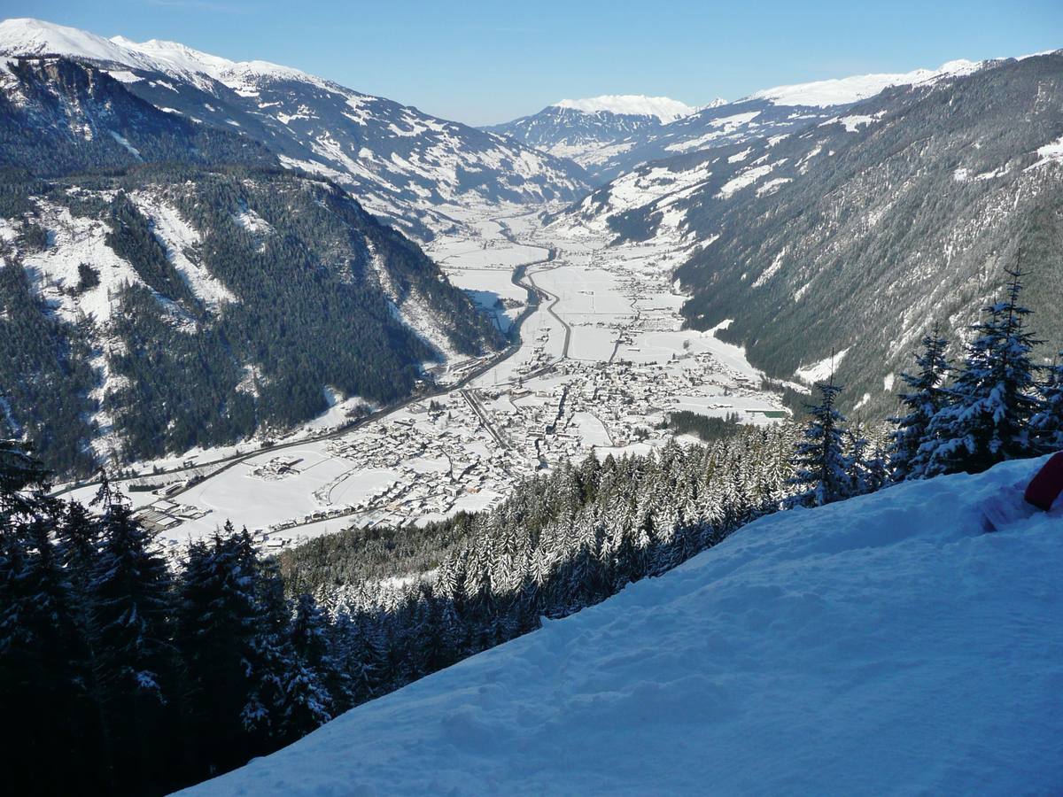 Mayrhofen's snow-covered rooftops filling the flat floor of the Zillertal, seen in sunshine from the Ahorn valley run above the town, with the river and valley road running north between forested slopes toward distant snowy peaks