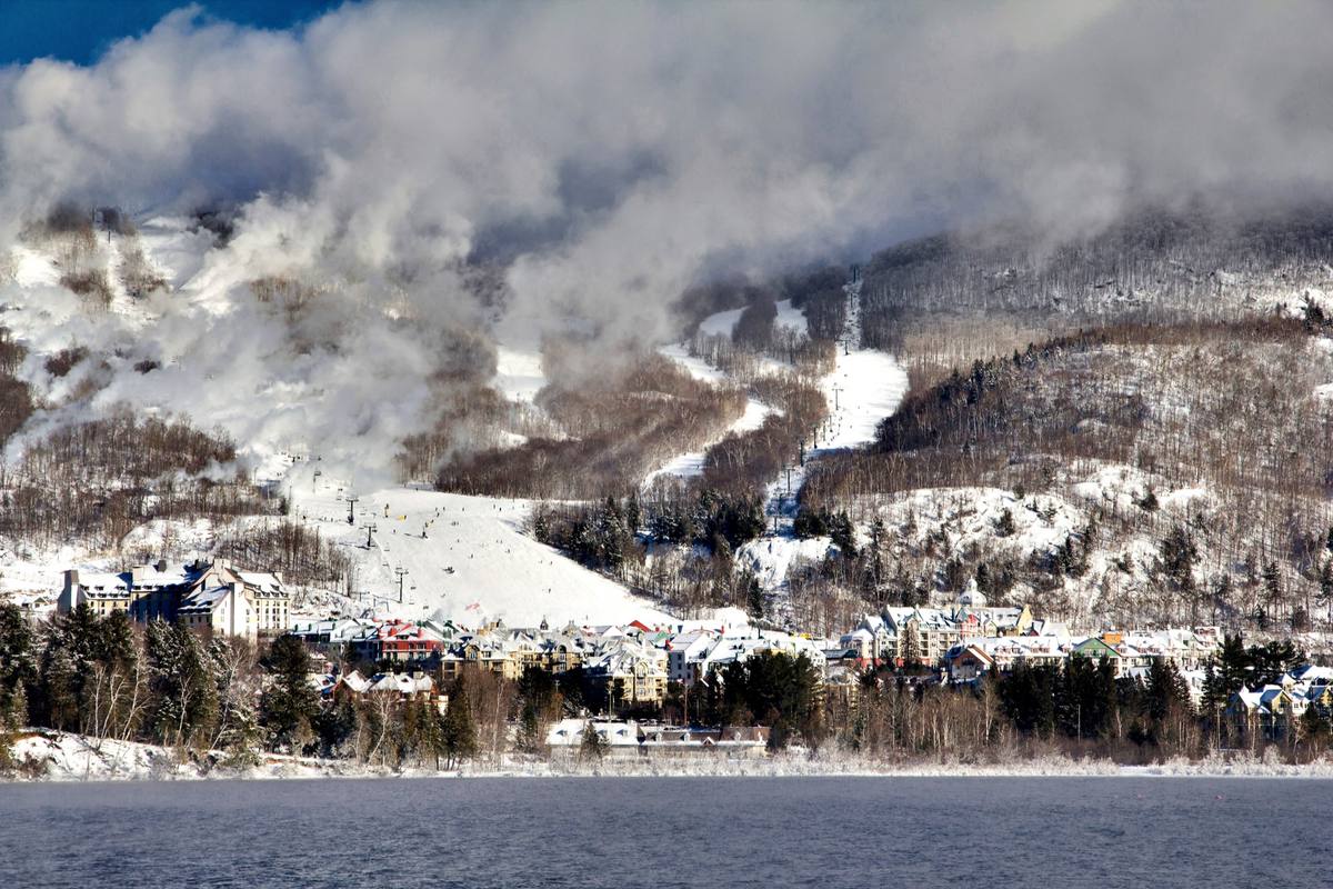 Mont-Tremblant's colorful pedestrian village along a frozen Lac Tremblant, with ski trails and snowmaking mist rising up the mountain behind