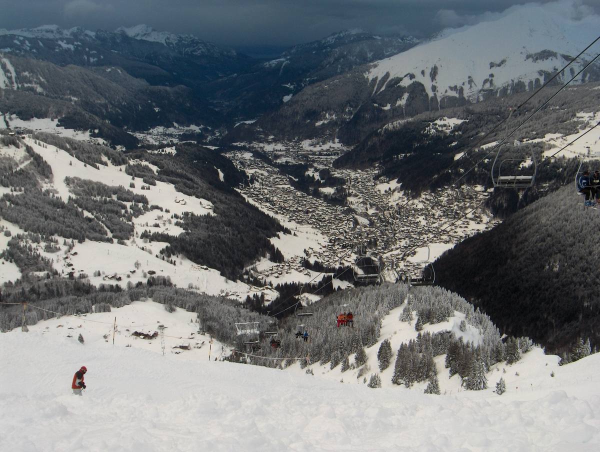 A snow-covered valley town of tightly packed chalet roofs seen from high on a piste above it, with a chairlift line running down the wooded slope in the foreground and dark storm cloud over the ridges beyond