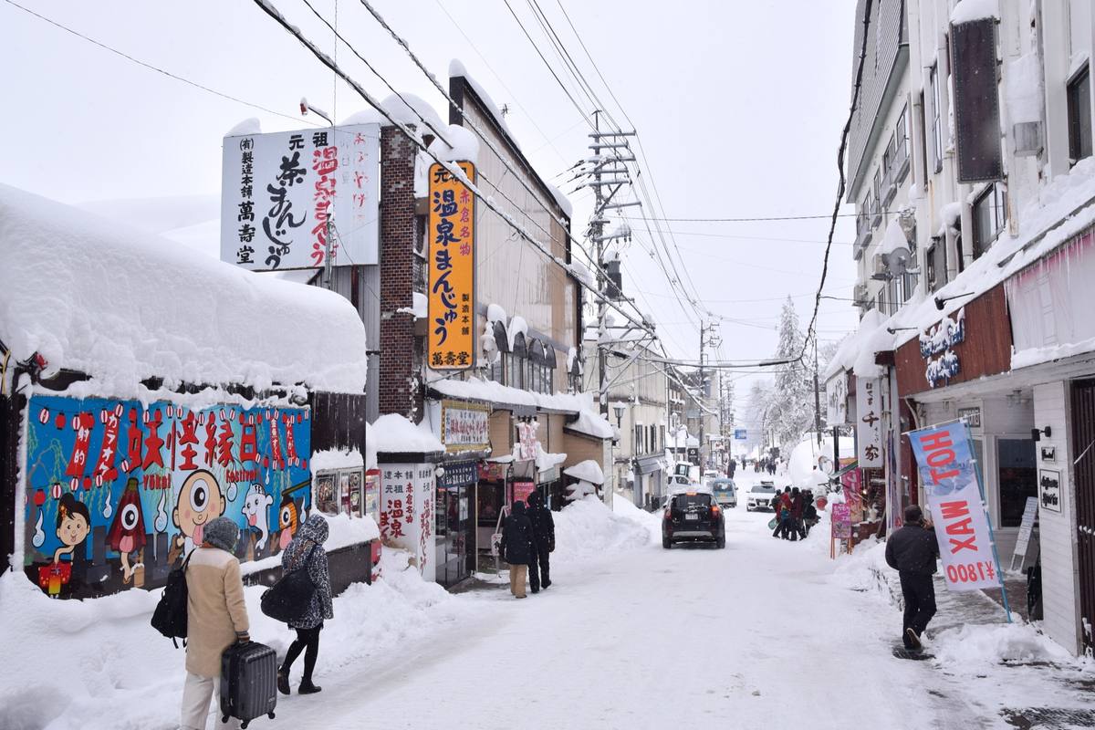 Akakura Onsen's main shopping street buried in deep snow, with thick snow caps on shop roofs and signage, a car navigating the cleared road, and pedestrians pulling luggage past storefronts