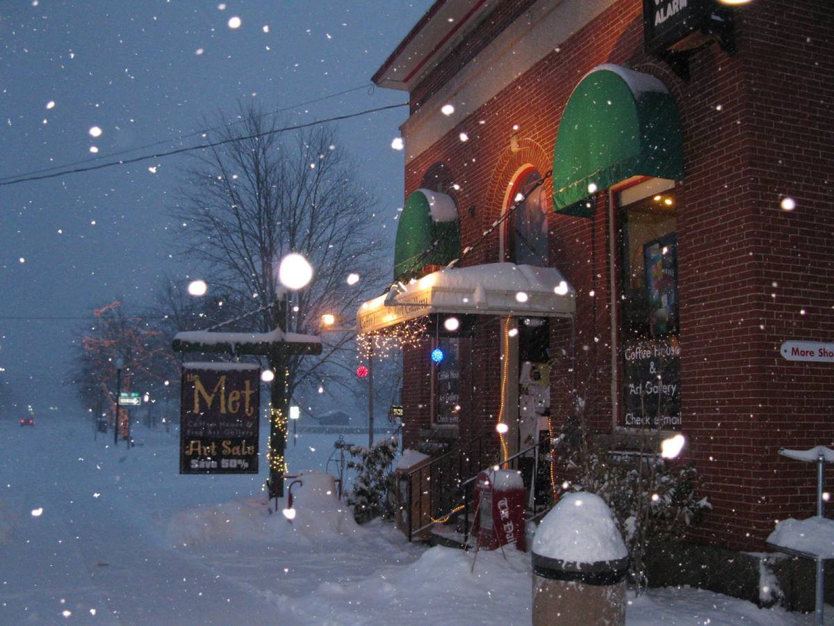 Snow falling at dusk on North Conway's main street, a lit brick storefront with green awnings and strings of lights above deep snowbanks