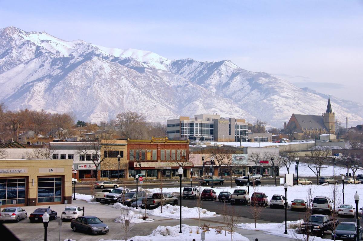 Snow-dusted brick storefronts in downtown Ogden with the spired St. Joseph's church at right and the snow-covered Wasatch Range rising directly behind the city under a hazy winter sky