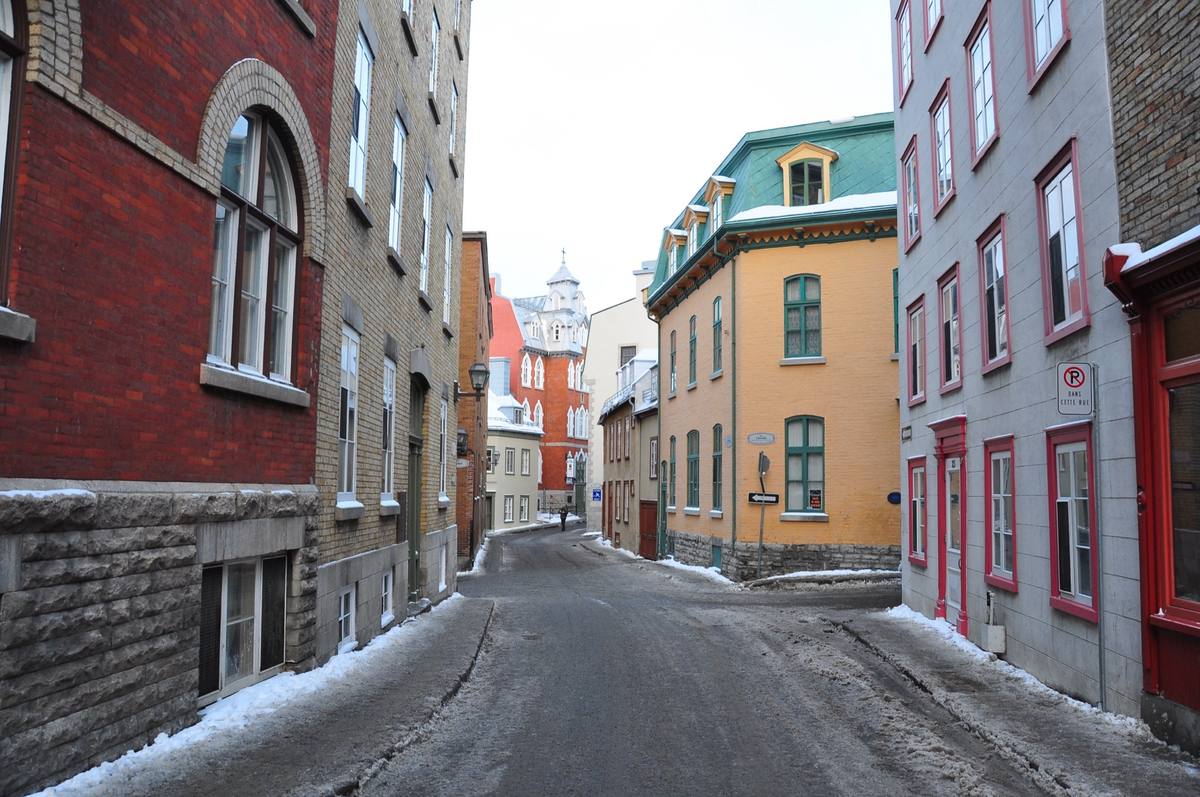 A snow-lined lane in Vieux-Québec, gray stone and red-brick row houses on either side, leading toward an ornate turreted building with a cross atop its spire