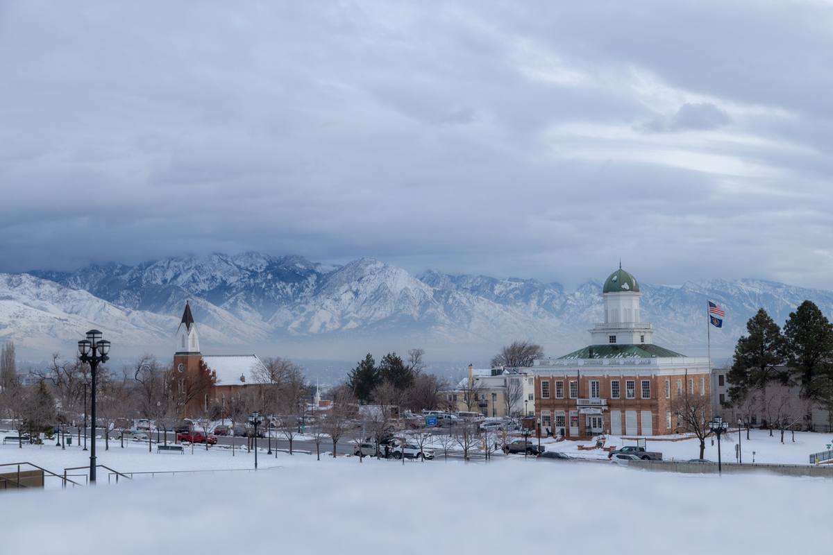 Snow-covered ground and bare trees around a domed red-brick civic building and a chapel spire on Capitol Hill, with a long snow-covered mountain range under heavy gray cloud beyond the valley