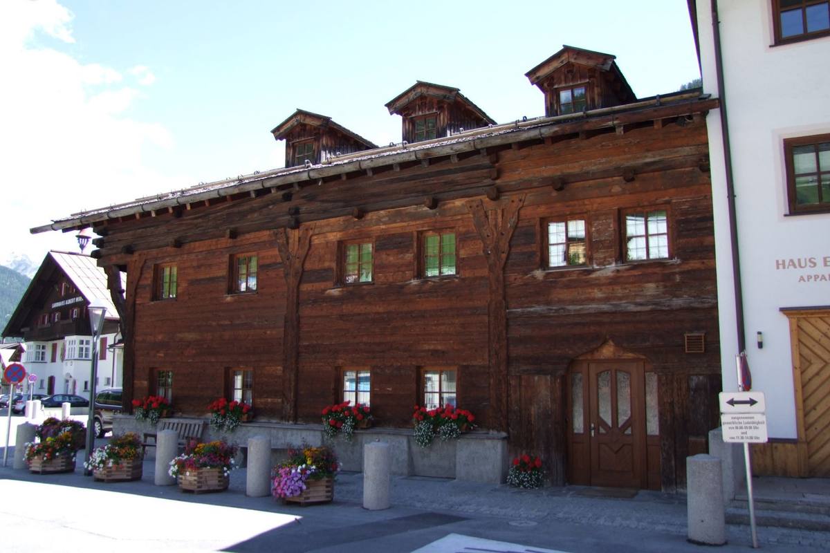The dark-timber Thöni Haus chalet with three roof dormers and geranium-filled window boxes on a village street in St. Anton am Arlberg, mountains visible behind