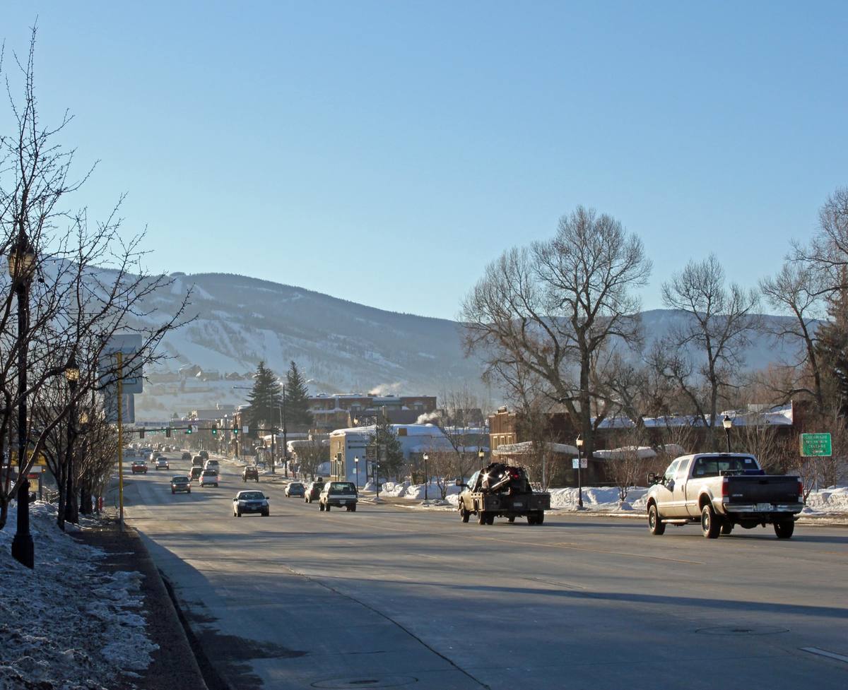 Traffic and pickup trucks on snow-banked Lincoln Avenue in low winter sun, brick storefronts and bare cottonwoods leading toward the snow-covered ski runs of Howelsen Hill