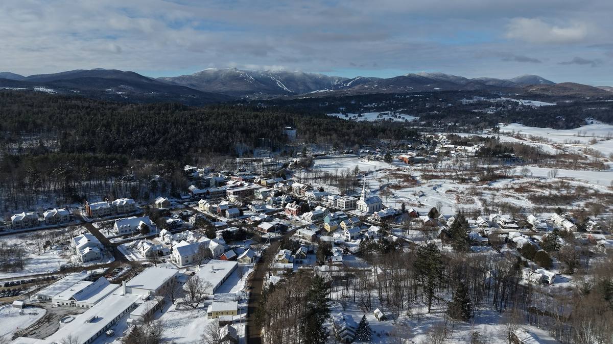 Stowe village under deep snow from the air, the white church spire at its center and the Mount Mansfield ridge with its ski trails on the skyline behind