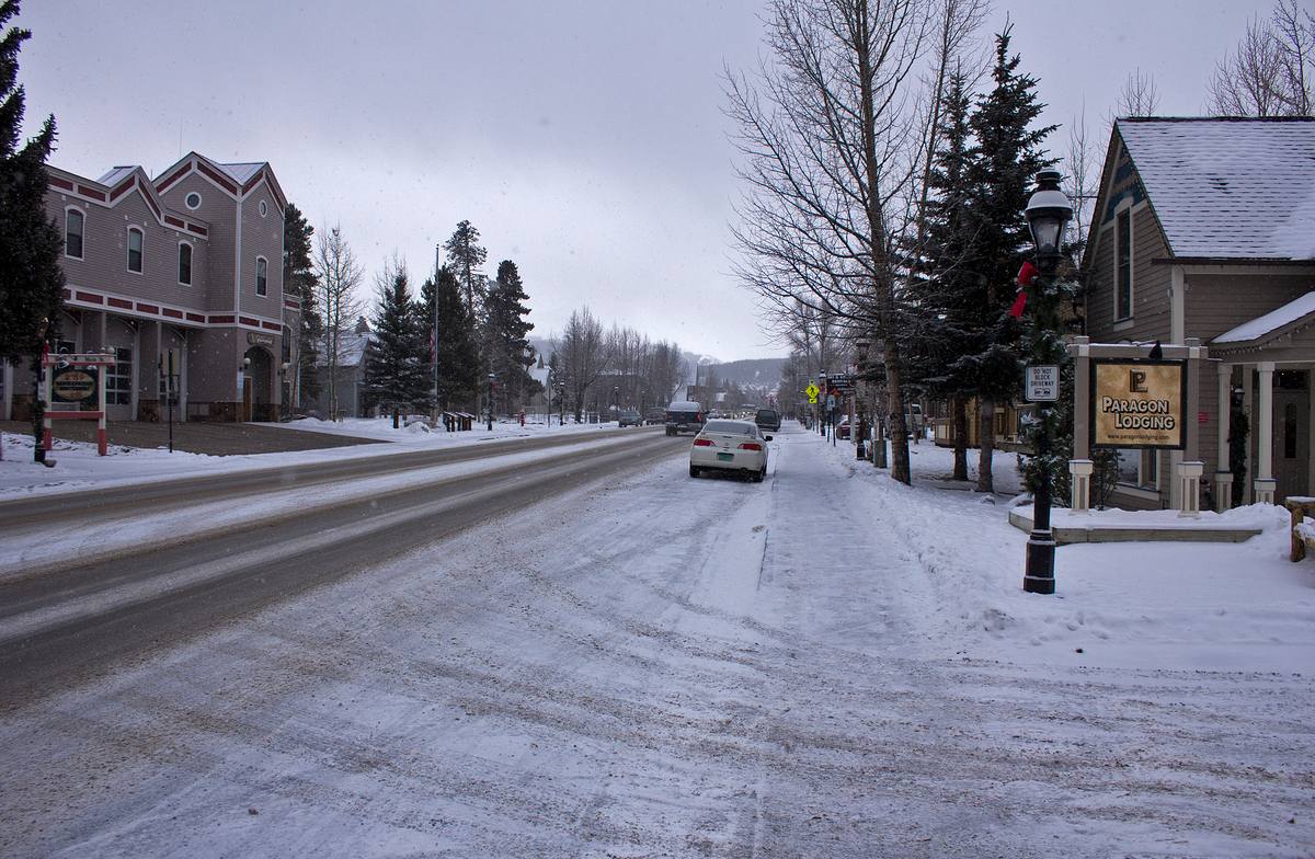 North Main Street in Breckenridge under falling snow, historic storefronts and the Paragon Lodging sign lining the road with a ski run visible on the mountainside at the end of the street