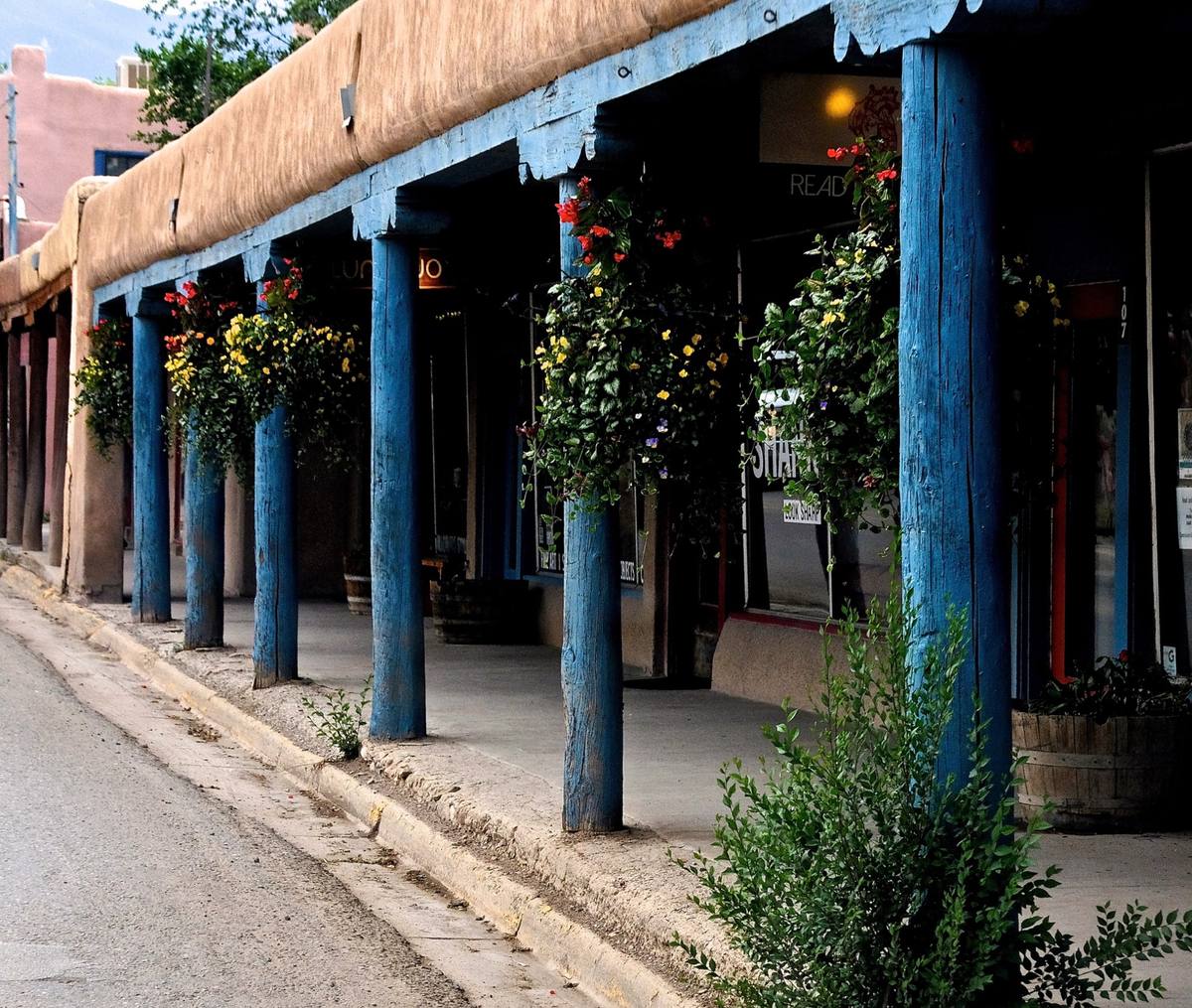 A shaded adobe portal on Taos Plaza with sky-blue support posts, hanging flower baskets, and gallery storefronts along the sidewalk