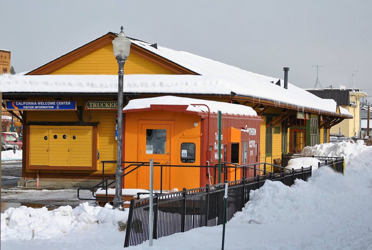 The snow-covered Truckee depot, marked TRUCKEE and California Welcome Center, with a bright orange rail car parked alongside, plowed snowbanks in front, and downtown storefronts visible behind it