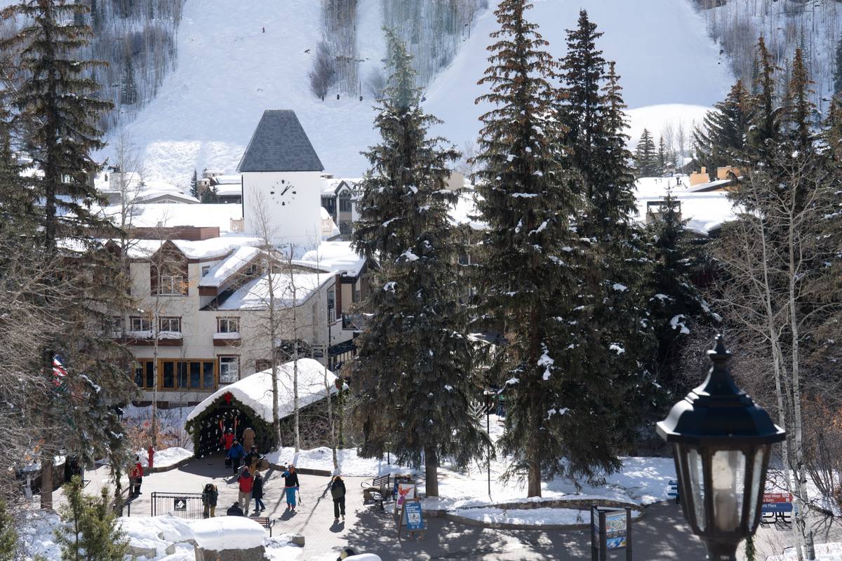 Skiers walking through deep snow past the wooden covered bridge in Vail Village, the clock tower and snow-laden roofs behind and groomed ski trails climbing the mountain above
