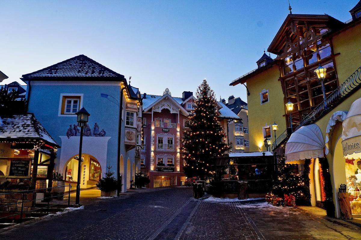 Ortisei's pedestrian square at blue hour in winter, snow on the rooftops and cobblestones, painted Tyrolean facades around a lit Christmas tree