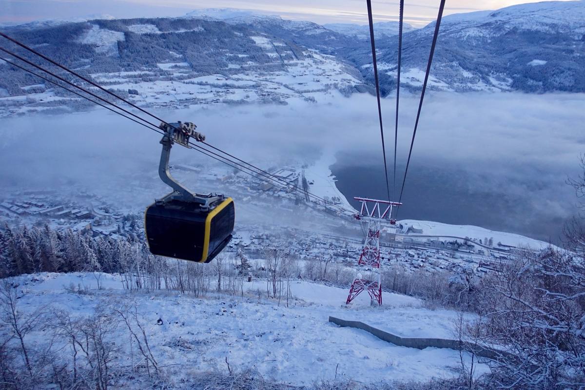 A gondola cabin descends toward snow-covered Voss, its cables and a support tower framing the town spread along fog-filled Vangsvatnet lake at winter dusk