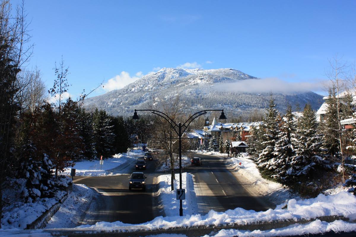 A plowed road curving into Whistler Village after fresh snow, snow-laden conifers and gabled timber buildings under a forested mountain slope and blue sky