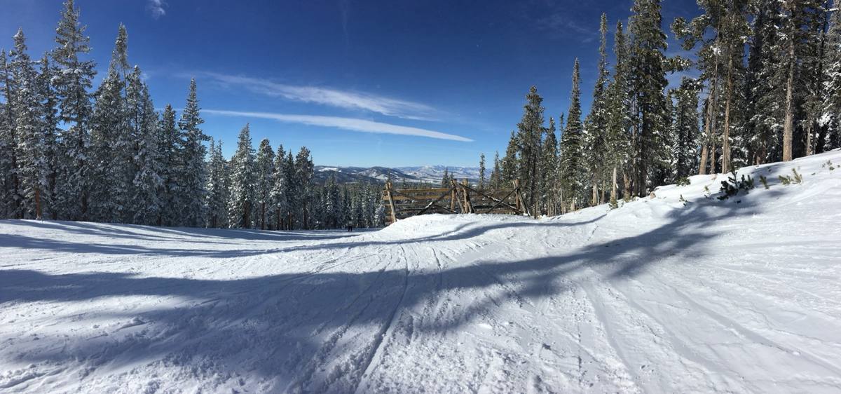 A freshly groomed ski run at Winter Park curving between snow-laden spruce trees under a deep blue sky, with a weathered timber snow fence and the peaks beyond the Fraser Valley on the horizon
