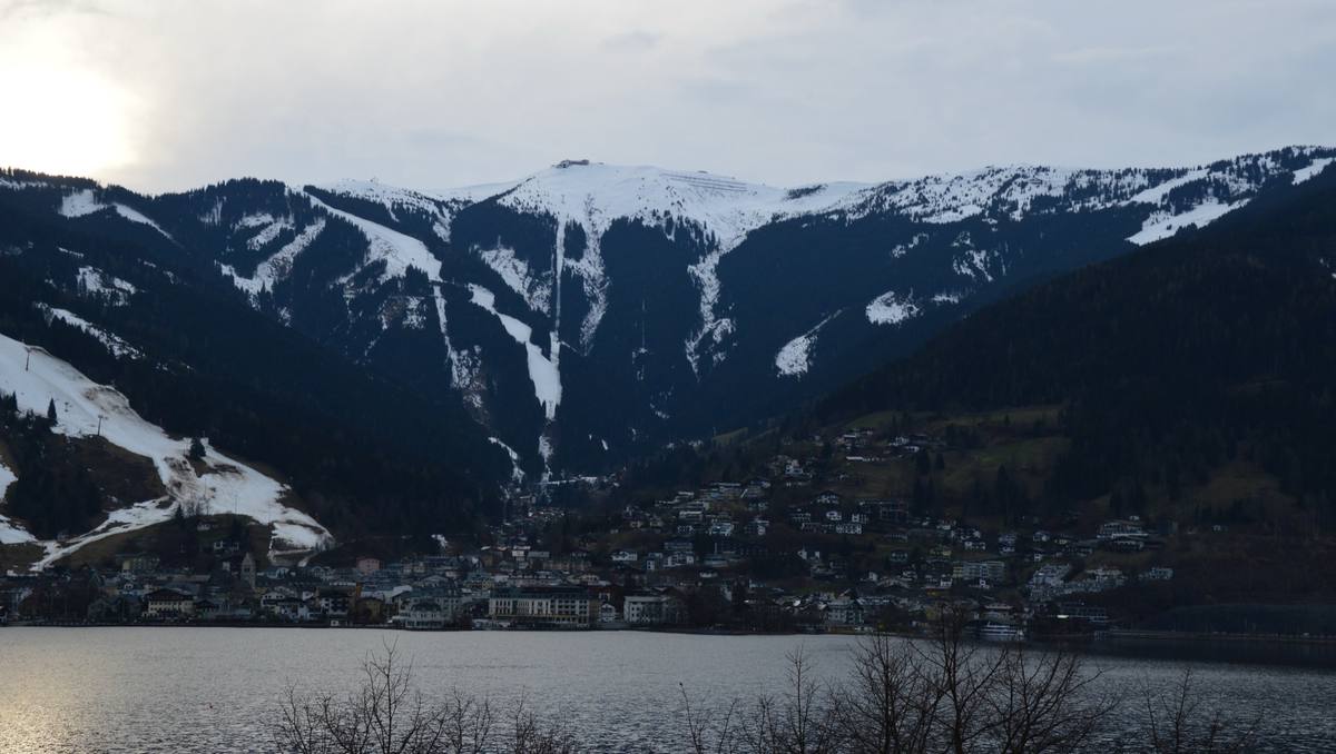 Zell am See's old town along the Zeller See lakeshore in winter, a church tower rising above the rooftops, with the snow-covered Schmittenhöhe and its ski runs behind